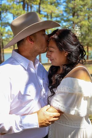 Groom in cowboy hat kisses bride on forehead at outdoor lakeside wedding.