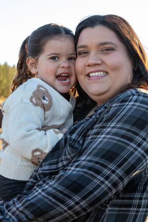Smiling mother holding her young daughter in a bear sweater outdoors in warm golden light.