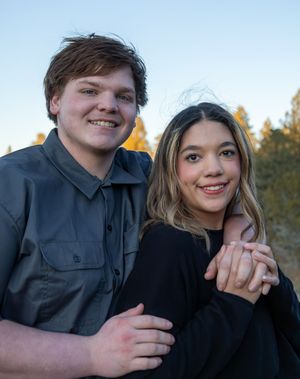 Smiling young couple posing closely together outdoors at dusk with trees in the background.