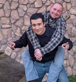 Two men laughing as one gives the other a piggyback ride against a stone wall.