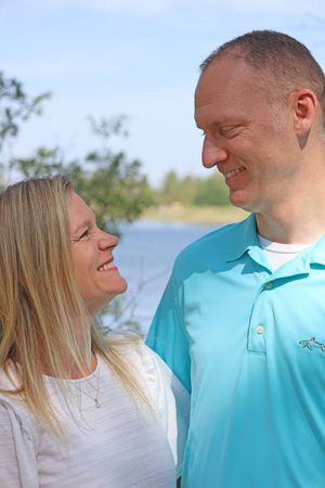 Smiling couple gazing at each other outdoors near a lake with trees and blue sky in the background.