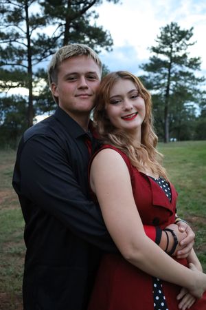 A young couple in formal attire embraces outdoors, she in red dress, he in black shirt, among pine trees.
