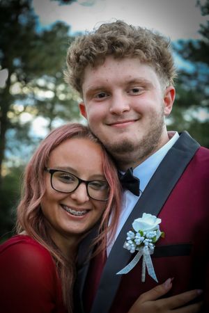 A smiling couple in prom attire poses together, he in burgundy tuxedo with boutonniere, she in red dress with pink hair.