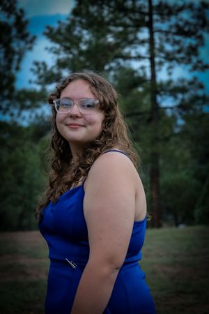 A teenage girl in a blue formal dress smiles over her shoulder outdoors among pine trees at dusk.