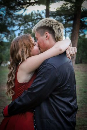 A young couple in formal prom attire shares a kiss outdoors surrounded by pine trees.