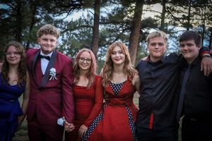 Six teenagers in formal prom attire pose together smiling outdoors among pine trees at sunset.