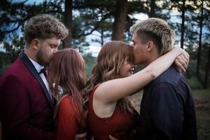 Two couples in formal prom attire share kisses in a romantic outdoor pine tree setting.
