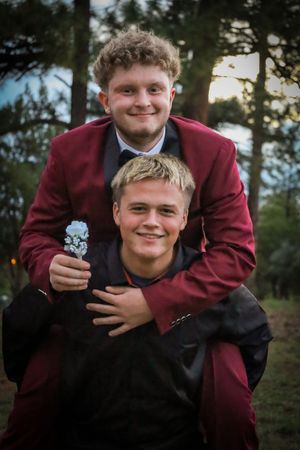 Two teenage boys in prom suits share a playful piggyback pose outdoors among pine trees.