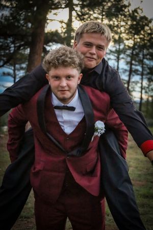 Two teenage boys in prom suits share a fun piggyback moment outdoors among pine trees at sunset.