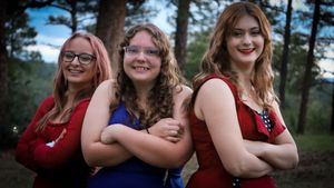 Three teenage girls in formal dresses pose with arms crossed back-to-back among pine trees.
