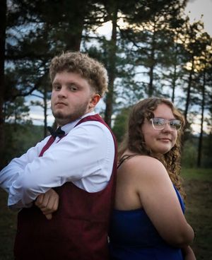 A teenage boy and girl in formal prom attire stand back-to-back outdoors among pine trees.