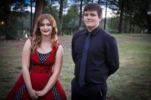 Teen couple dressed formally in red polka dot dress and black suit posing outdoors among pine trees.