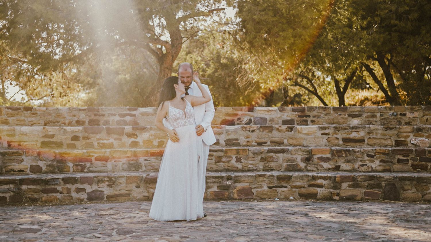 A couple stands together in elegant attire against a stone wall with sunlight streaming through autumn trees.