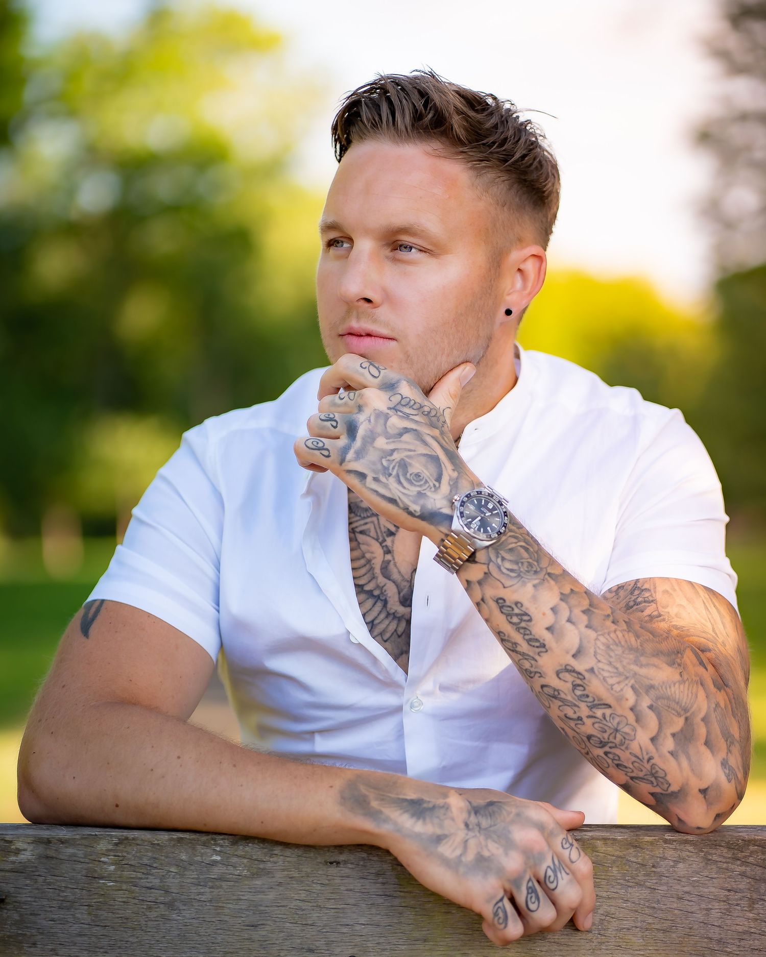A person in a white shirt with intricate tattoo sleeves poses thoughtfully outdoors.