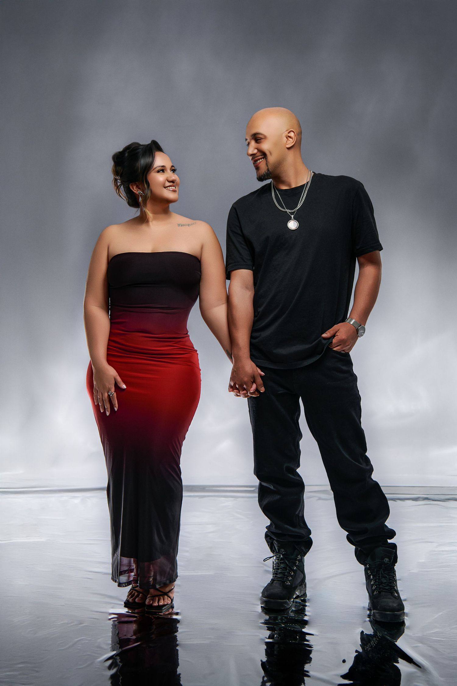 A romantic couple poses together on a reflective surface, with her in a red dress and him in black casual wear.