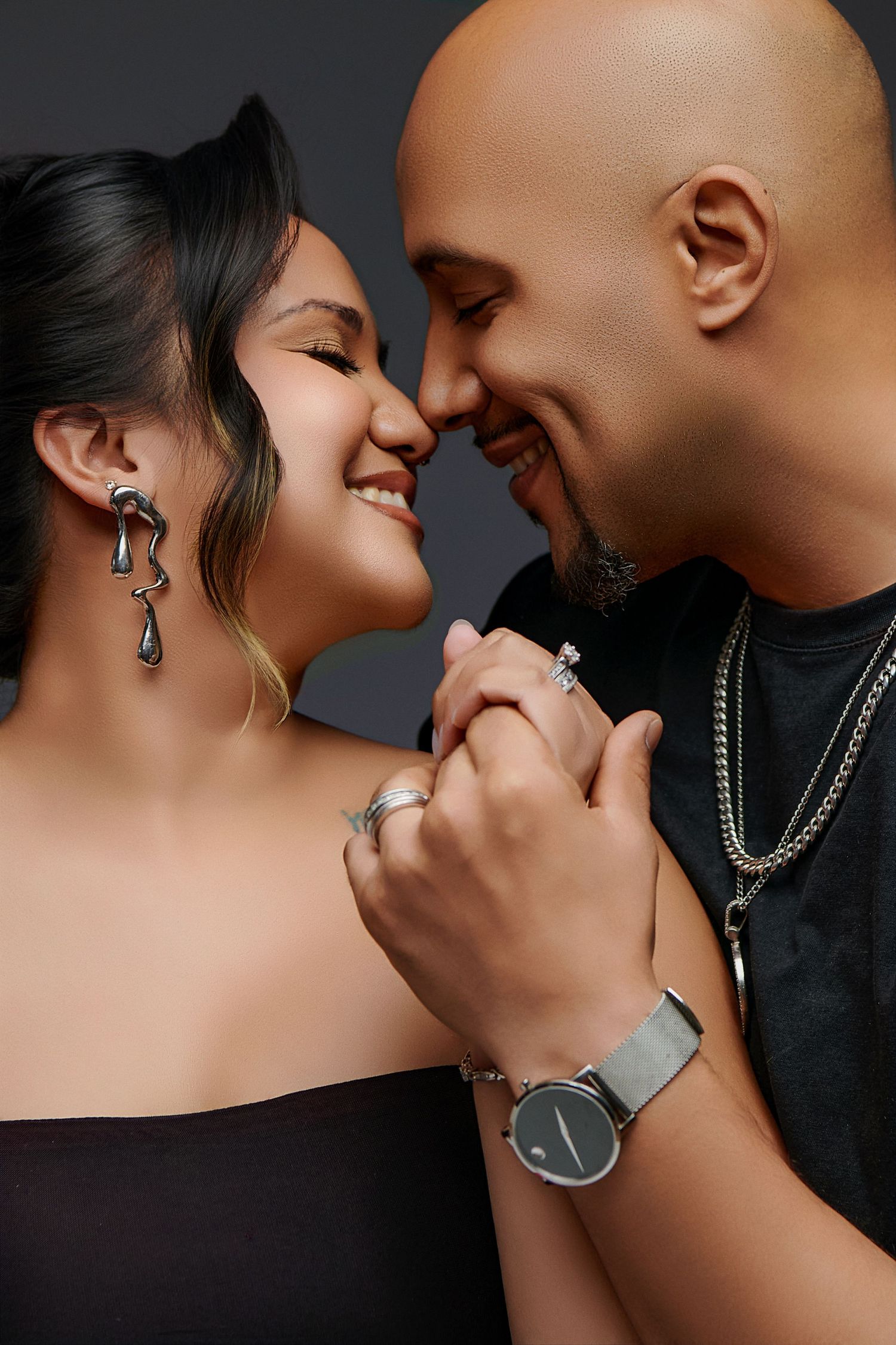Intimate couple in black attire share a romantic moment while holding hands, featuring elegant jewelry and a wristwatch.