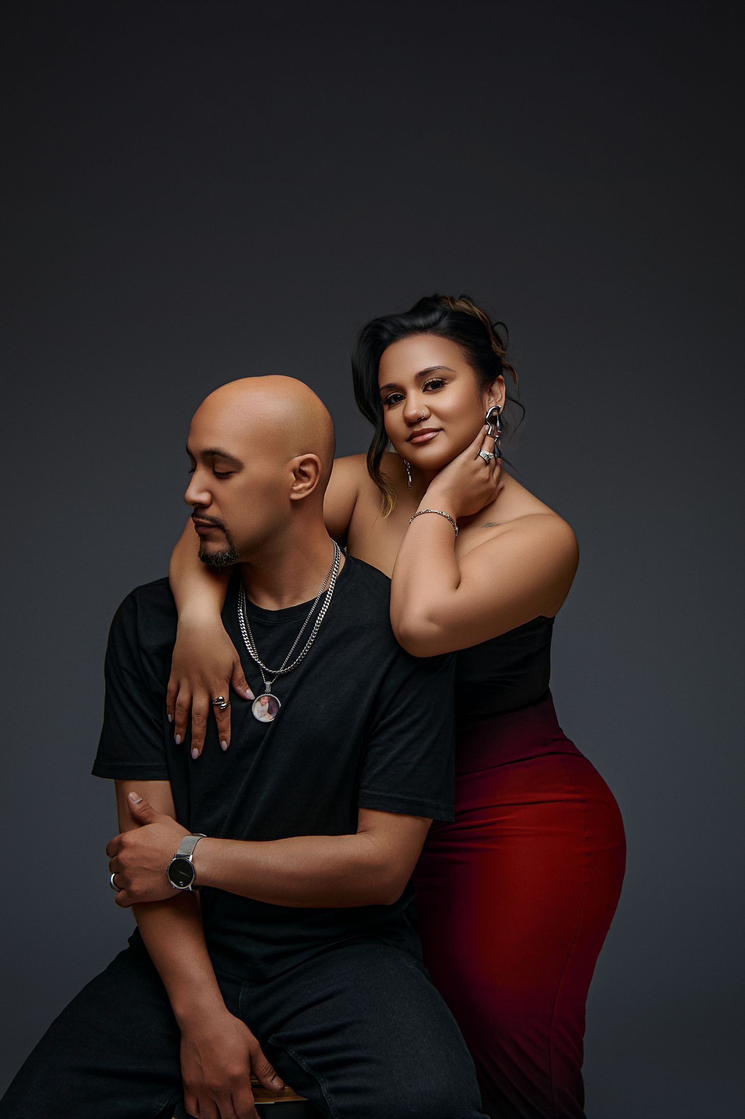 Studio portrait of a couple posing intimately against dark background, with her in red dress and him in black attire.