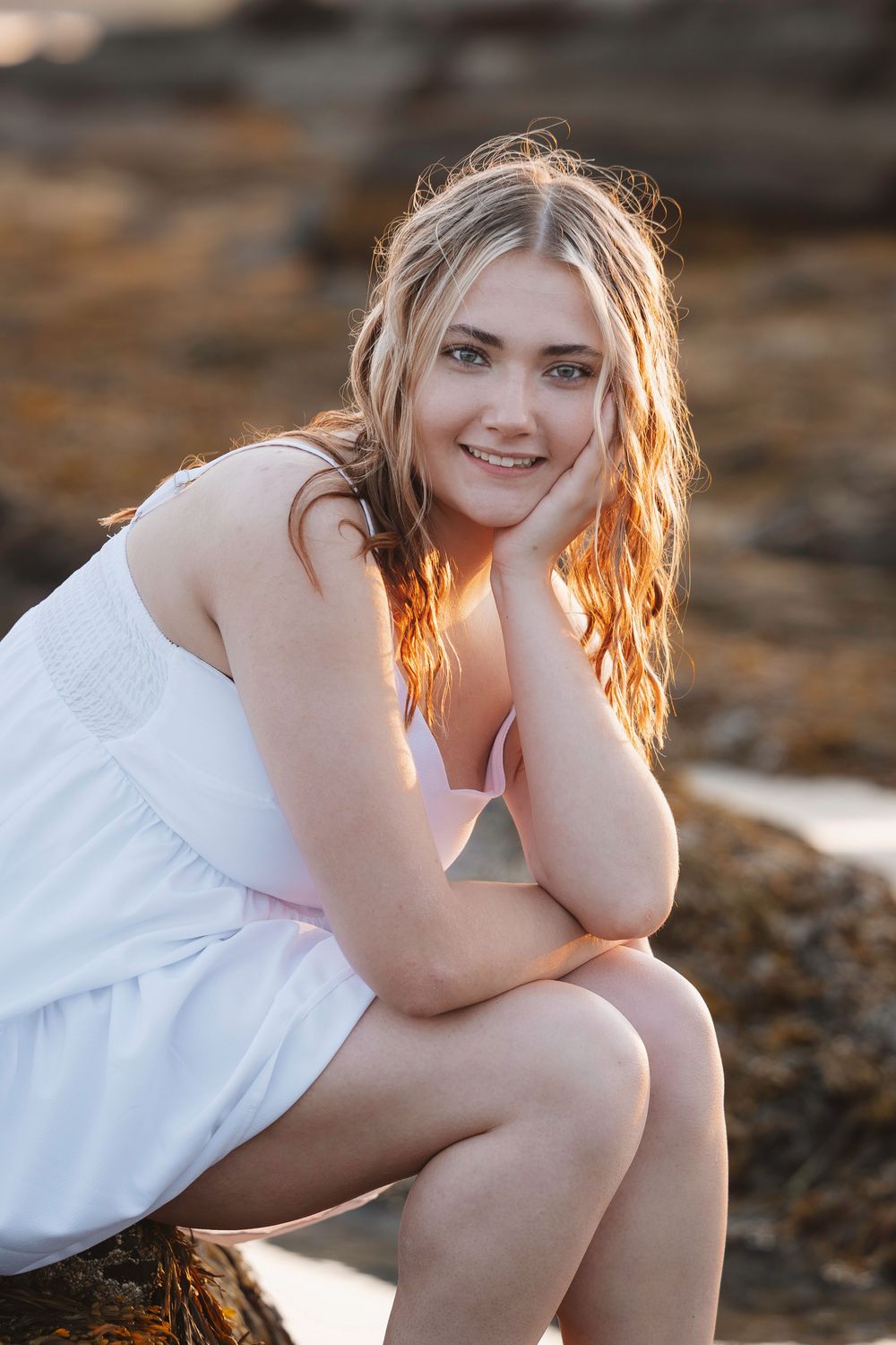 A woman in a white dress sits on rocks at golden hour, with sunlight illuminating her hair from behind.