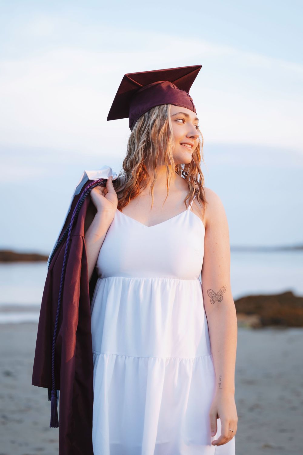 Graduate wearing white sundress and burgundy graduation gown stands smiling near ocean shoreline during seaside photoshoot.
