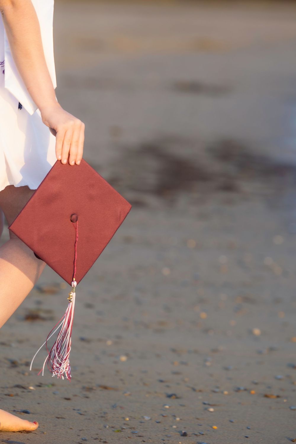 A person holding a red graduation cap with tassel while walking barefoot on a sandy beach.