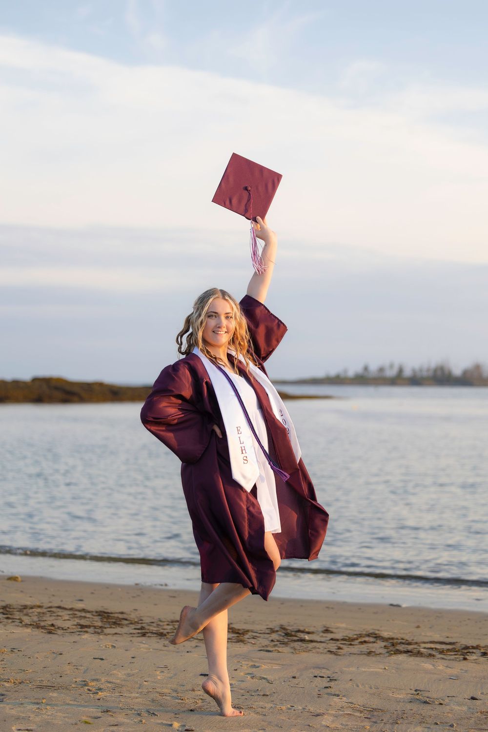 A graduate in maroon cap and gown celebrates on a beach at sunset, holding their diploma high in the air.