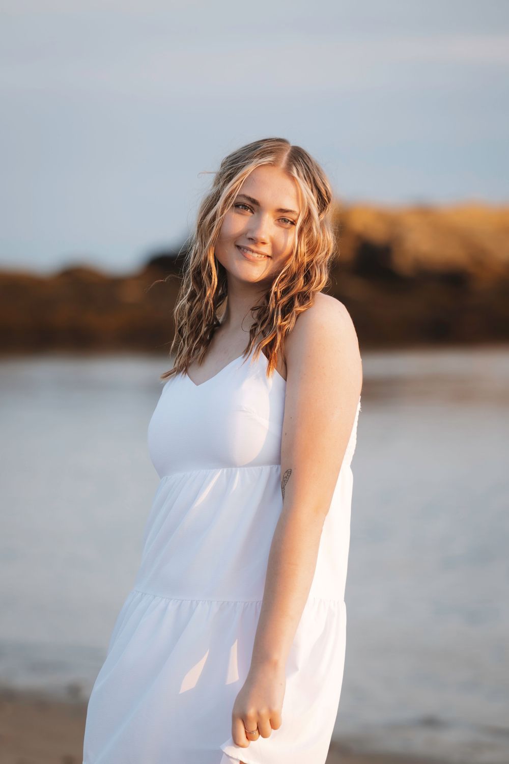 A person in a flowing white dress stands on a beach at sunset with water and golden hills in the background.