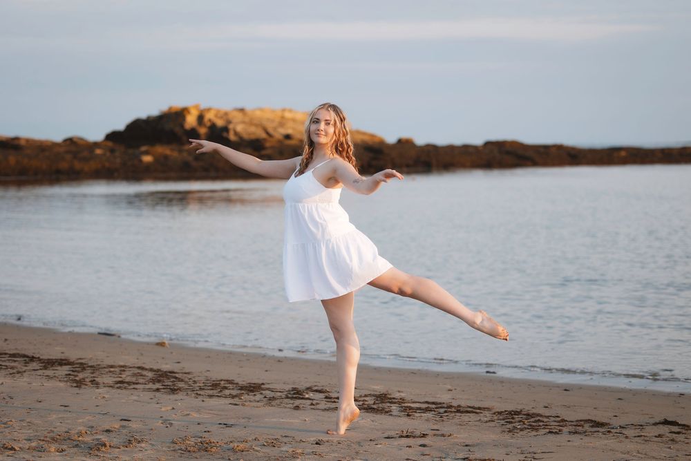 Dancer in flowing white dress performs graceful pose on beach at sunset by calm water.