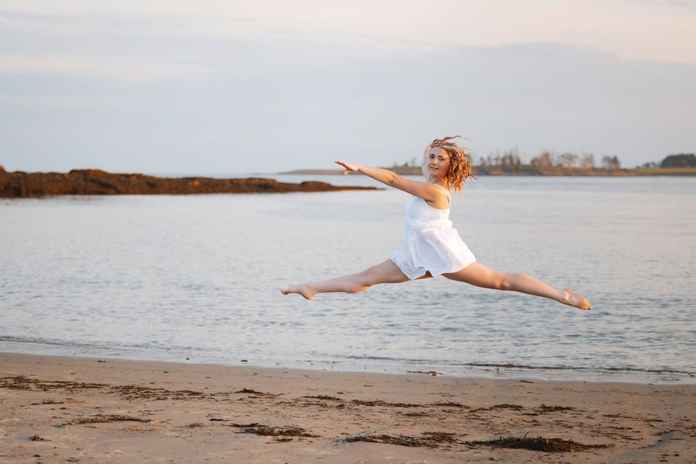 A person joyfully leaps and dances along the sandy beach at sunset, creating silhouettes against the calm ocean water.