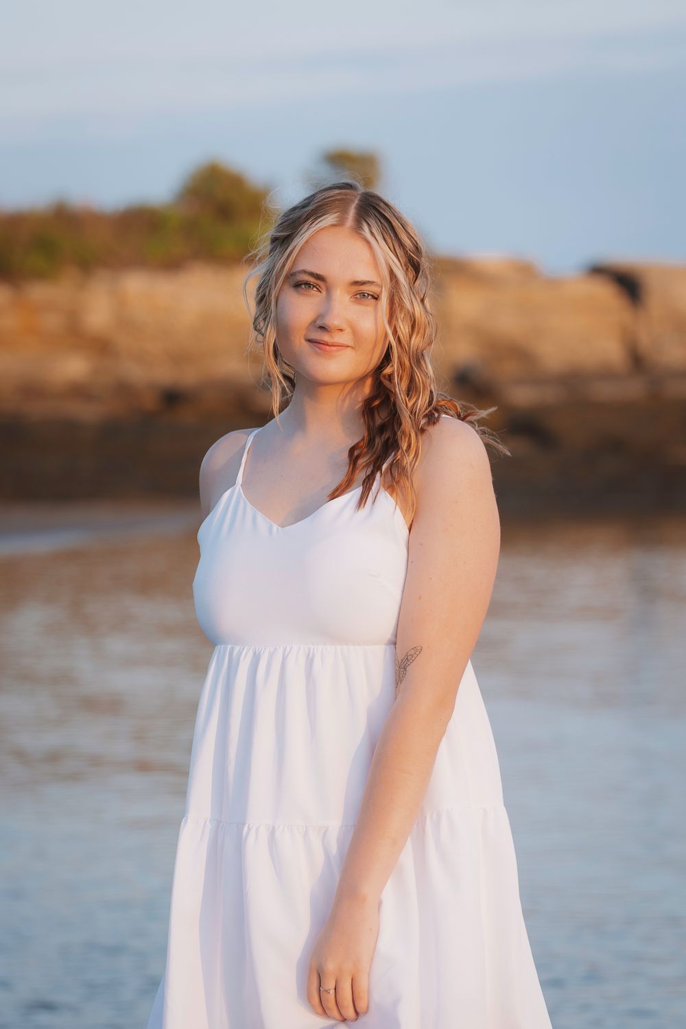 Woman in white flowing sundress poses along rocky coastline at sunset with ocean and cliffs in background.
