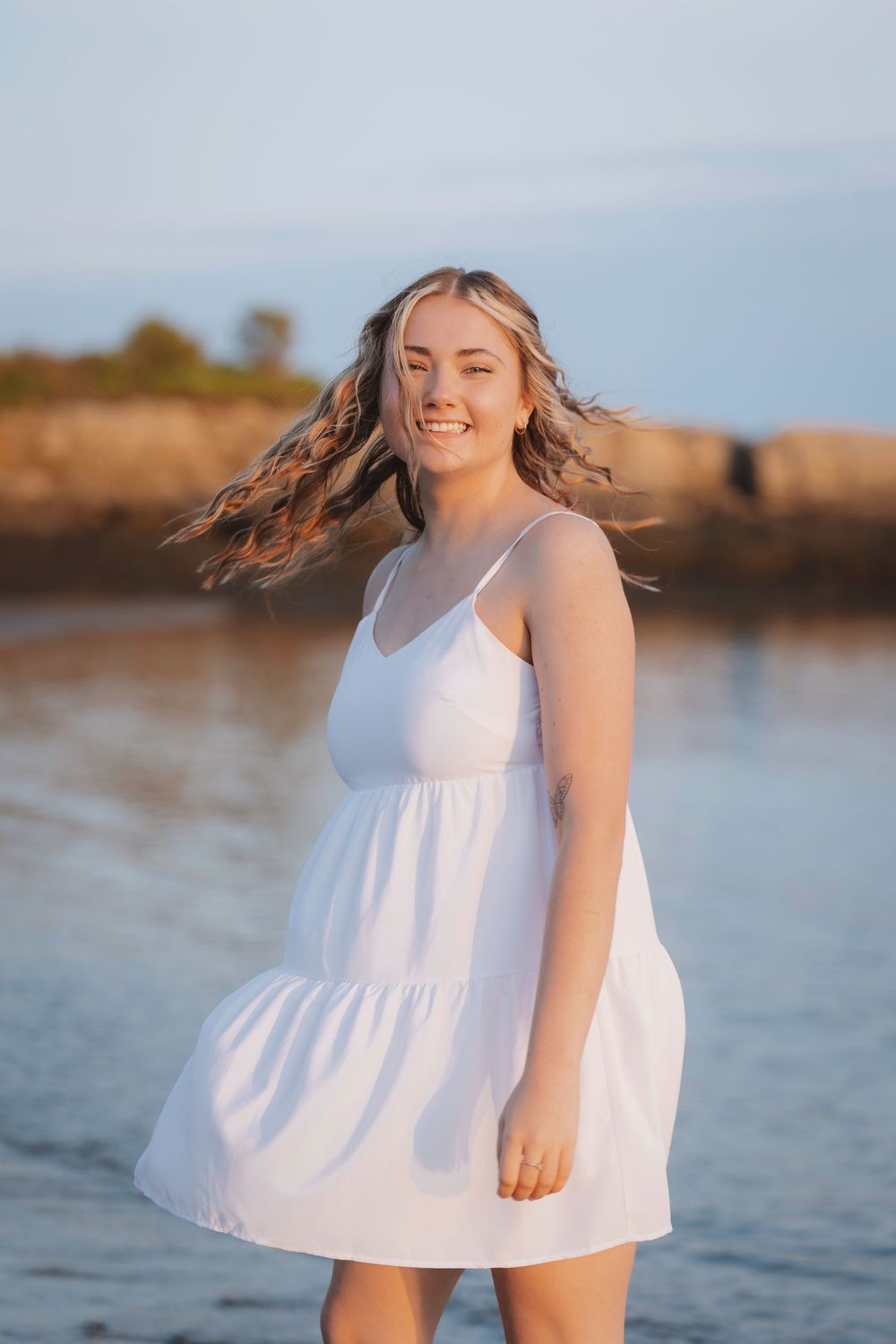 A young person in a flowing white dress twirls joyfully by the ocean during golden hour.