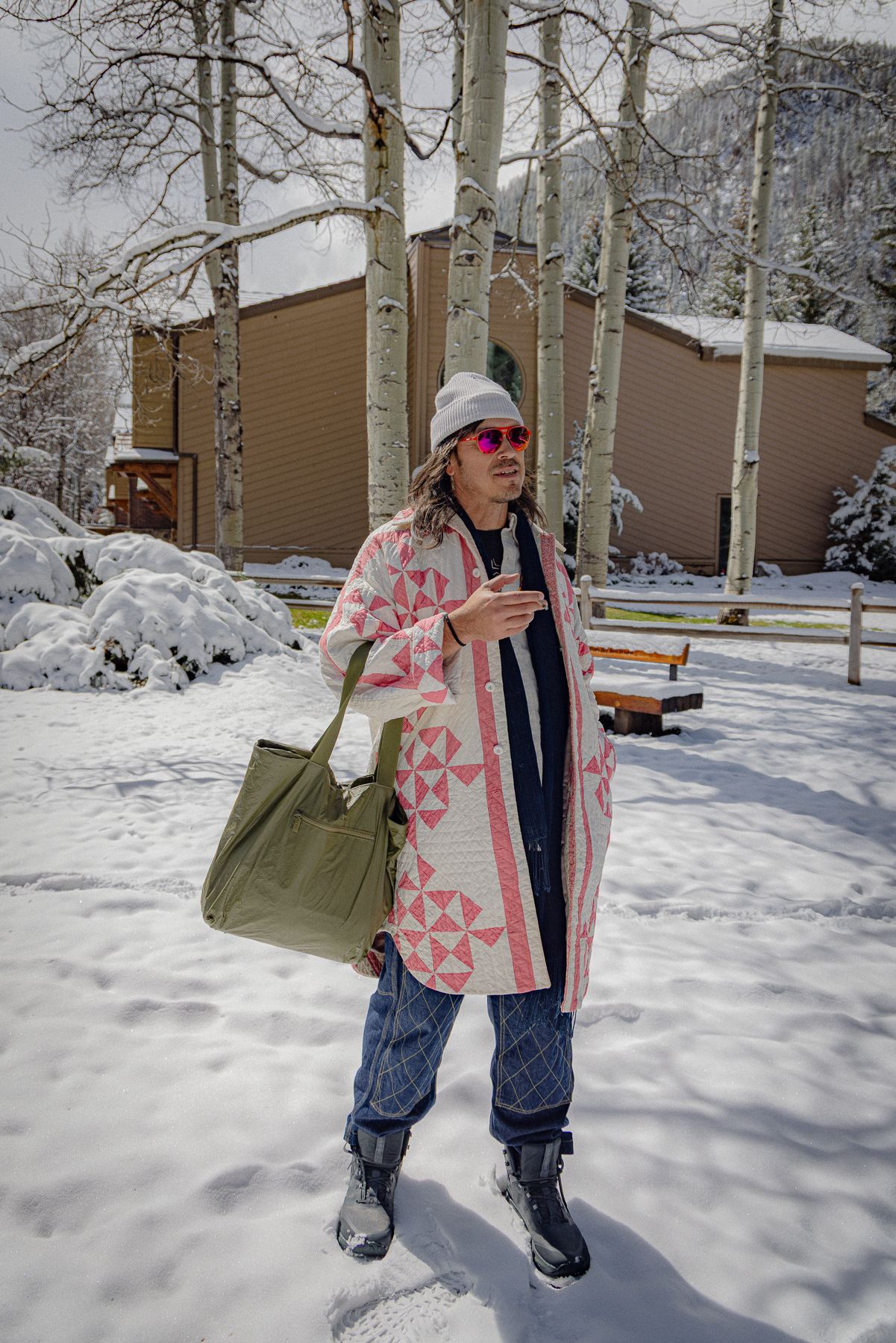 Someone in a pink patterned coat and blue jeans stands in a snowy winter scene with aspen trees and buildings in background.