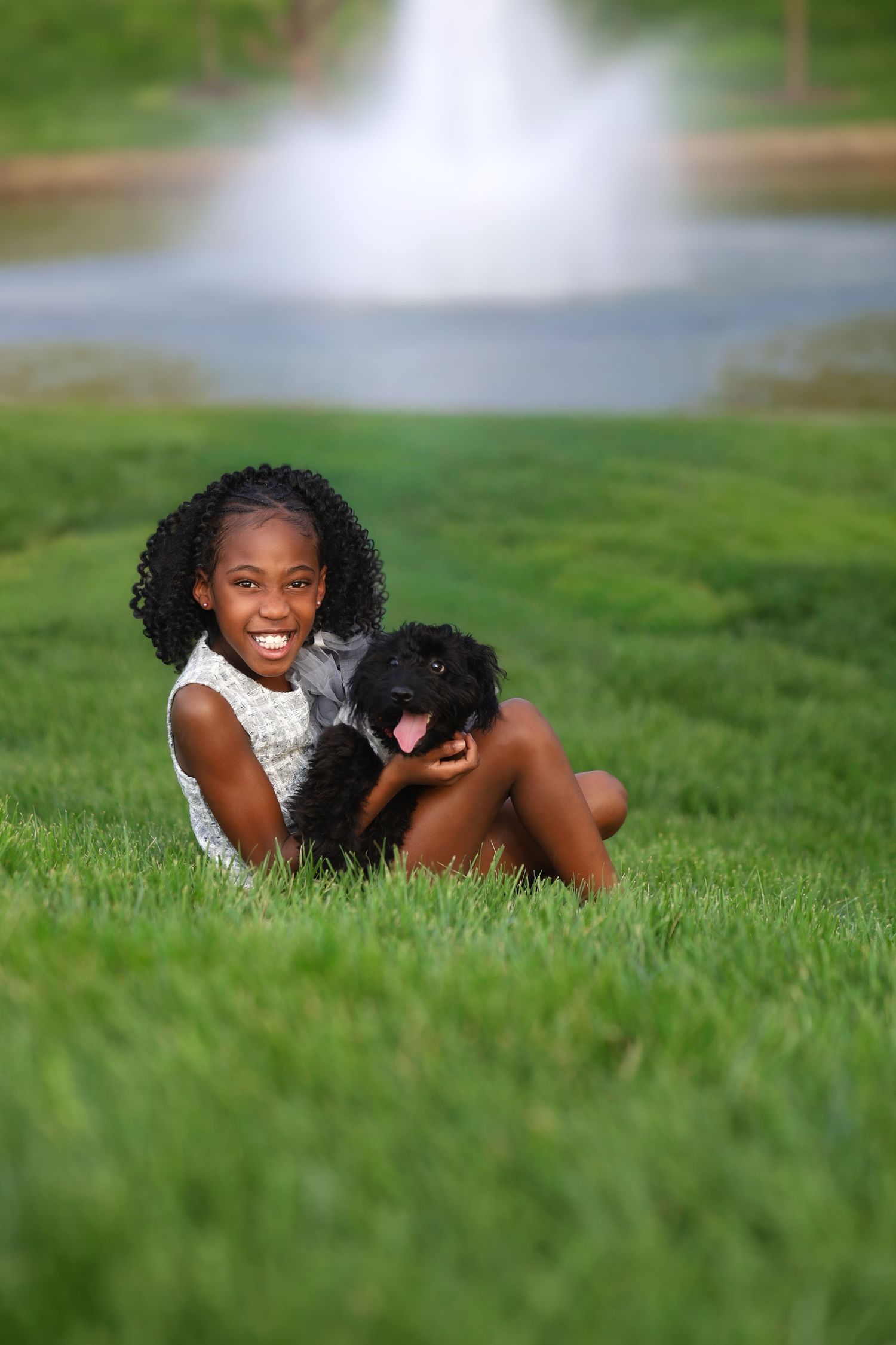 Smiling young Black girl sits cross-legged on green grass holding a black poodle puppy near a fountain.