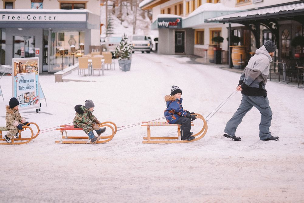 A parent pulls children on wooden sleds through a snowy shopping area lined with stores and cafes.