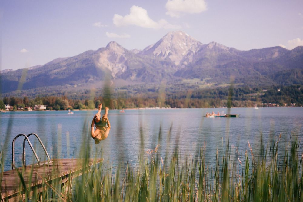 Person diving into peaceful mountain lake surrounded by tall peaks and reeds.