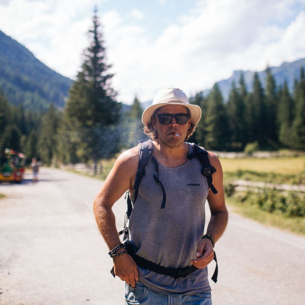 Hiker in hat and tank top on mountain trail with evergreen forest backdrop.