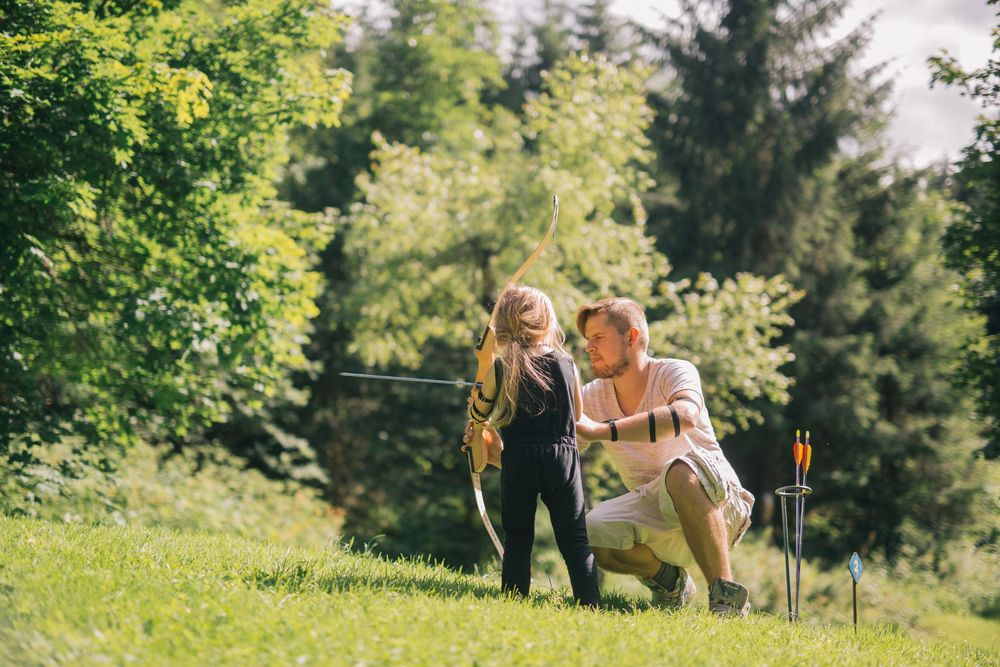 Two people explore a sunny grassy hillside with evergreen trees in the background on a bright summer day.