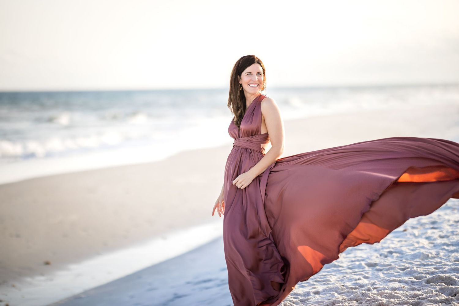 A flowing burgundy dress billows in the ocean breeze as someone poses on a sunlit beach at golden hour.