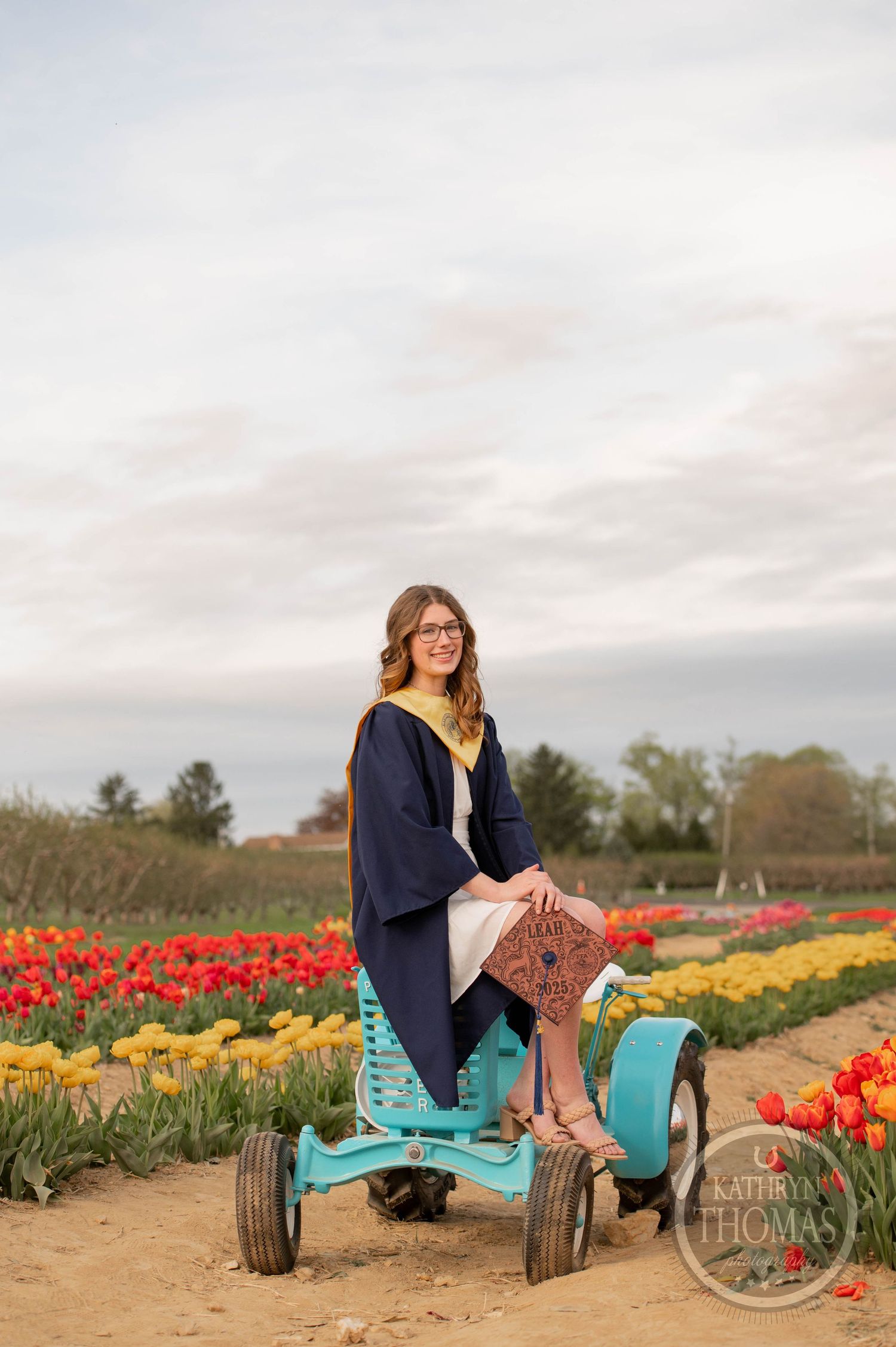 Graduate poses among vibrant blooming tulip fields with metal grain silos in the background under cloudy skies.