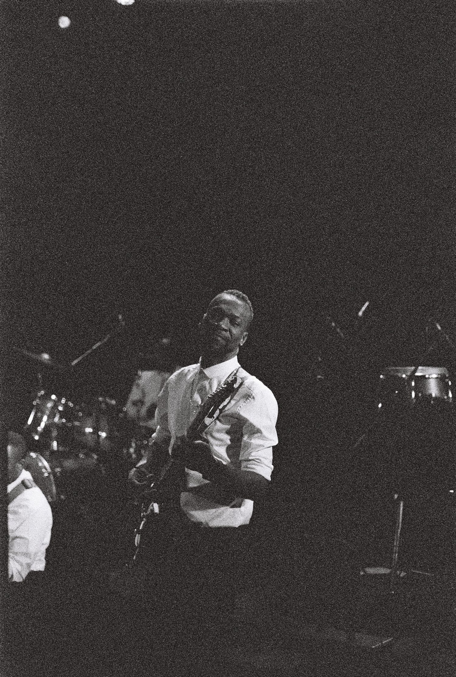 Black and white photo of a performer playing guitar on stage with drums in background.