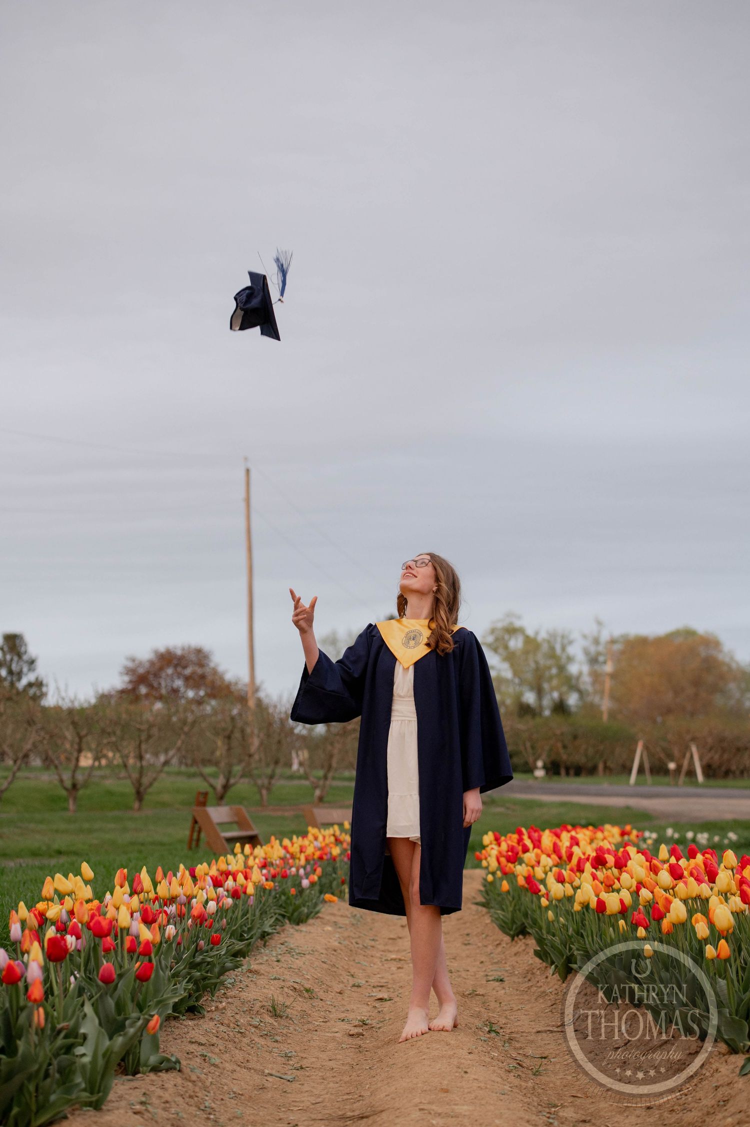 Graduate throws cap in celebration while standing on dirt path between vibrant orange and yellow tulip fields under cloudy sky.