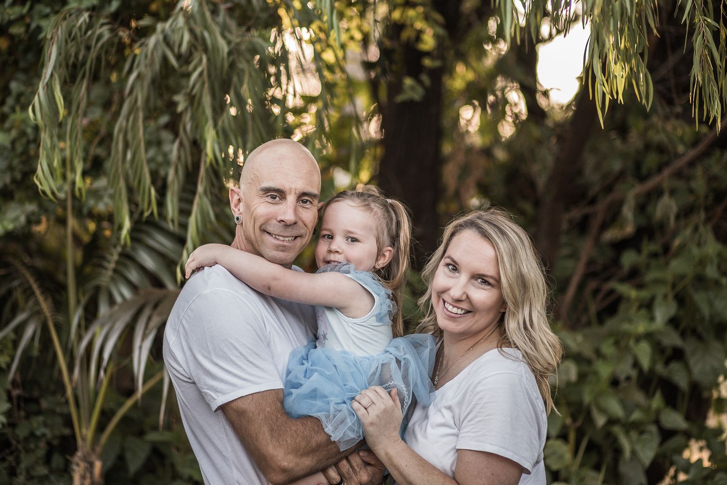 A family poses together outdoors in white shirts against a backdrop of lush tropical foliage and palm trees.