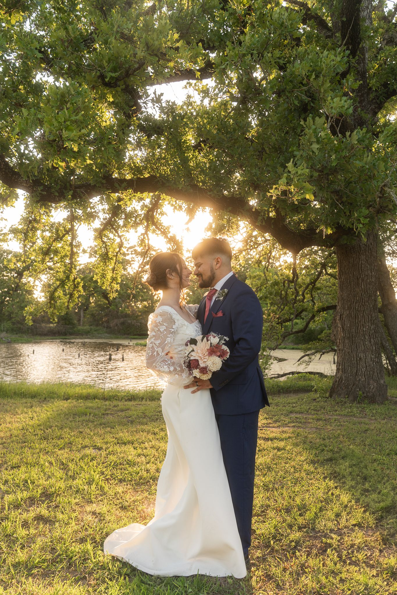Couple shares romantic sunset moment by lake with golden light filtering through trees and illuminating white wedding dress.