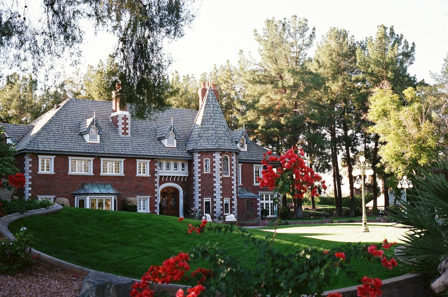 Large brick mansion with Tudor-style architecture surrounded by red flowers and green trees on a manicured lawn.