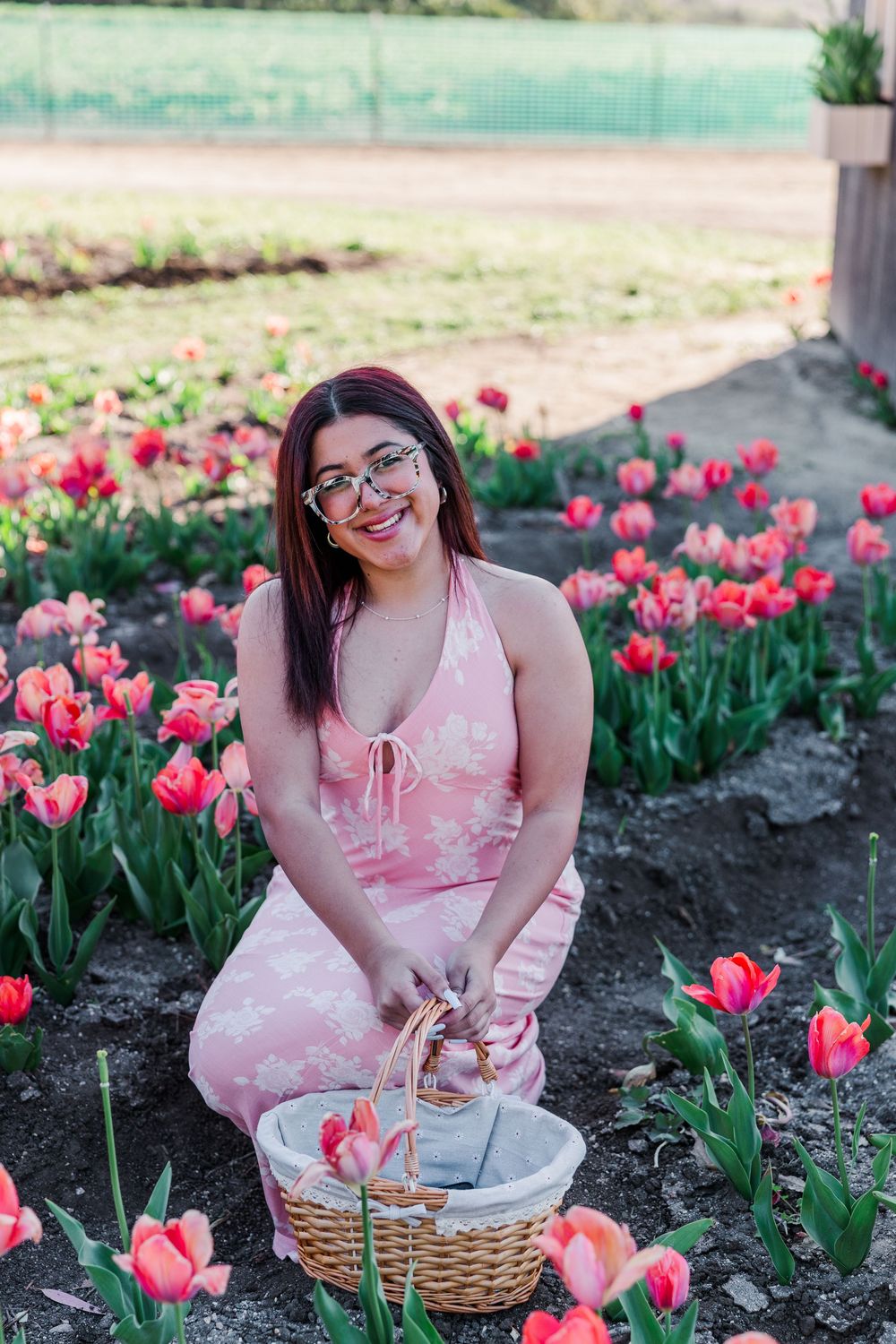 A person in a pink dress sits among blooming pink tulips in a garden with a wicker basket beside them.