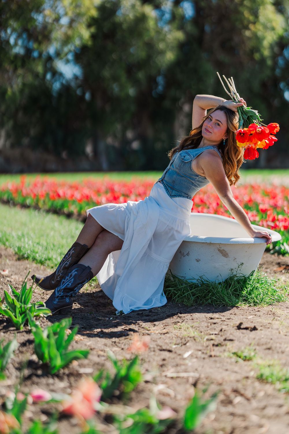 A woman in a blue top and white dress poses gracefully among blooming tulips in a garden setting on a sunny spring day.