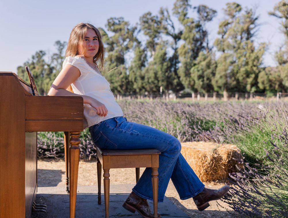 A person sits casually at a piano outdoors near hay bales and tall trees in a rustic country setting.