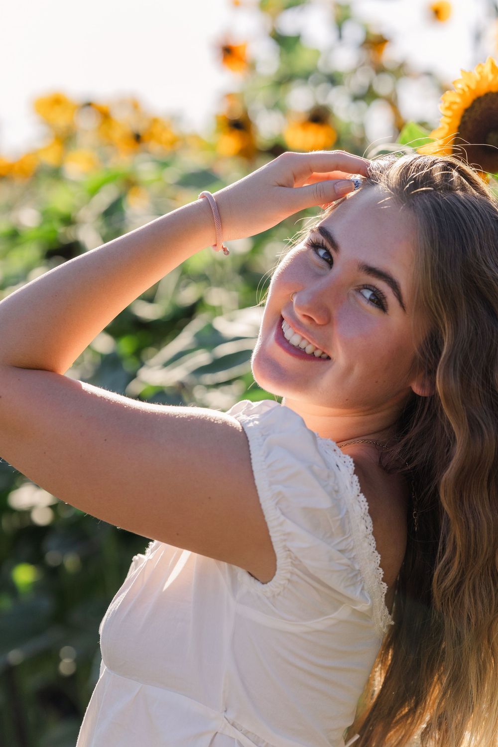 Woman in white dress smiles while shielding eyes from sun in sunflower garden during golden hour.