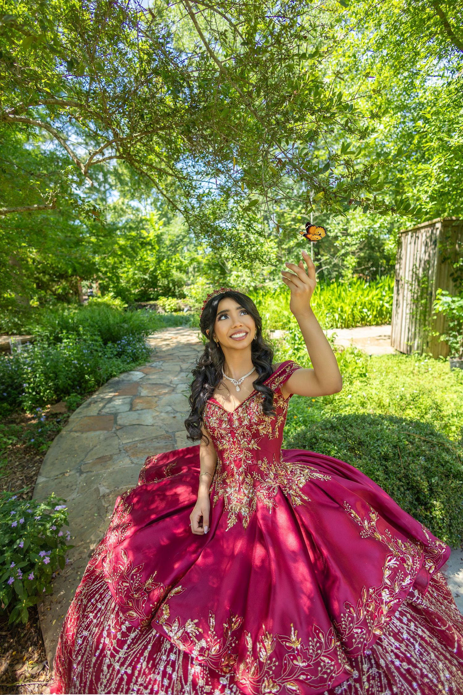 A burgundy quinceañera dress with silver embellishments sparkles against a lush garden backdrop.