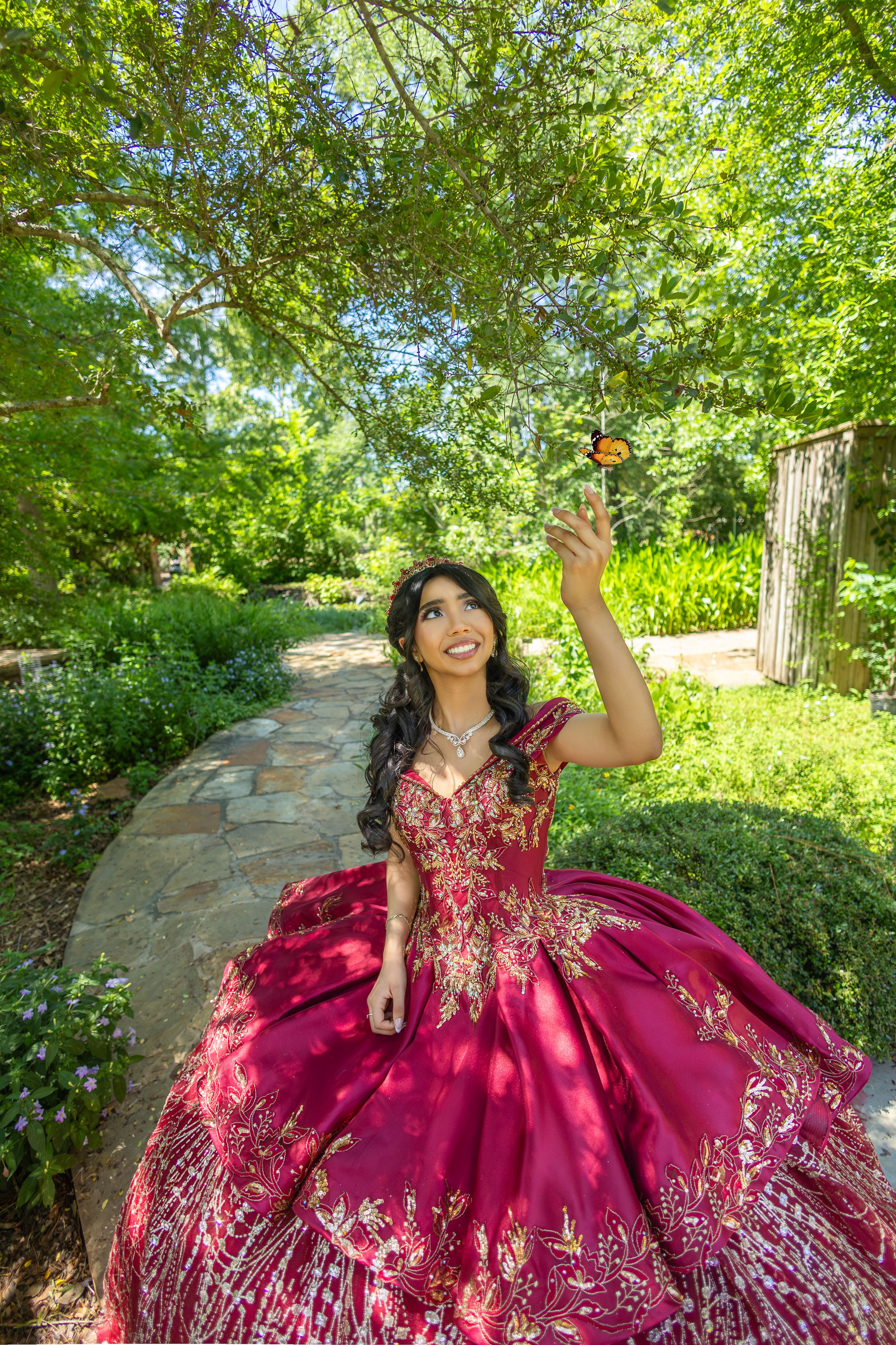 A burgundy quinceañera dress with silver embellishments sparkles against a lush garden backdrop.