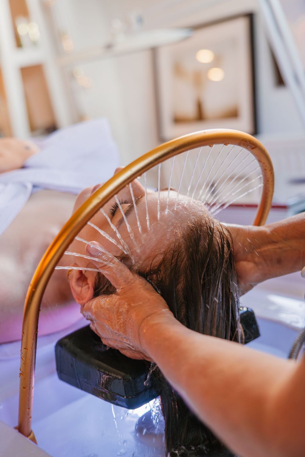 Hairdresser washing and rinsing dark hair at salon sink with curved faucet and white basin.